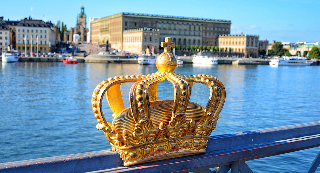 Bild på en kungakrona avbildad på en bro, med Stockholms slott i bakgrunden. Foto: Santi Blanco/Pixabay.